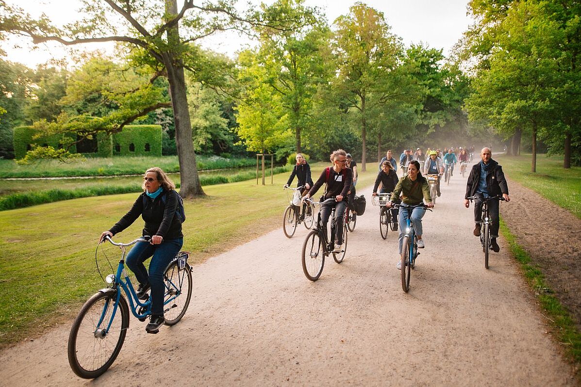 Exkursion Eine Gruppe Radfahrender auf einem breiten Parkweg, umgeben von Bäumen und Rasenflächen.