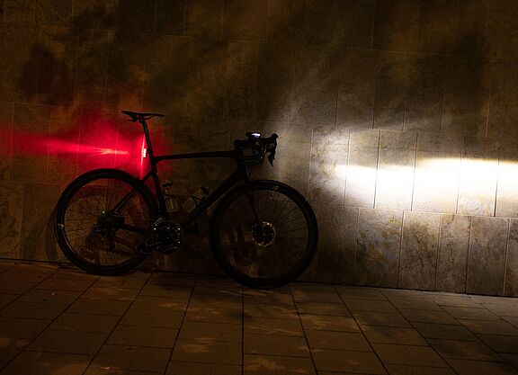 Sigma Aufmacher Ein Fahrrad steht in der Dunkelheit an einer Wand, mit am Lenker montiertem, eingeschalteten Scheinwerfer und an der Sattelstütze montierten, leuchtenden Rücklicht.