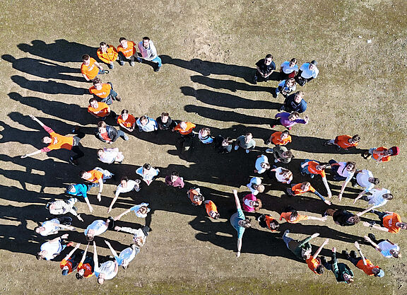 Menschen bilden ein Fahrrad Gruppe von Menschen steht auf einem Platz und bildet gemeinsam die Form eines Fahrrads. Die Personen tragen teils orangefarbene, teils weiße oder bunte Oberteile. Das Bild wurde aus der Vogelperspektive aufgenommen.