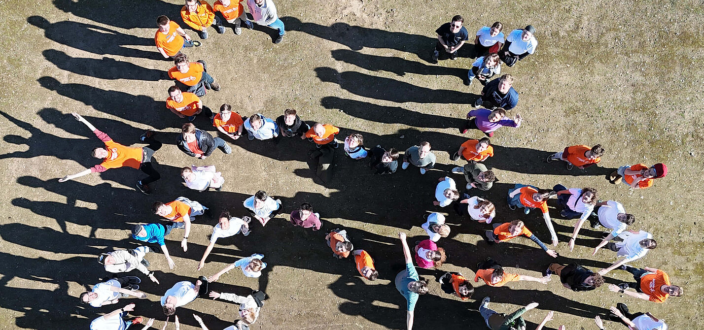 Menschen bilden ein Fahrrad Gruppe von Menschen steht auf einem Platz und bildet gemeinsam die Form eines Fahrrads. Die Personen tragen teils orangefarbene, teils weiße oder bunte Oberteile. Das Bild wurde aus der Vogelperspektive aufgenommen.
