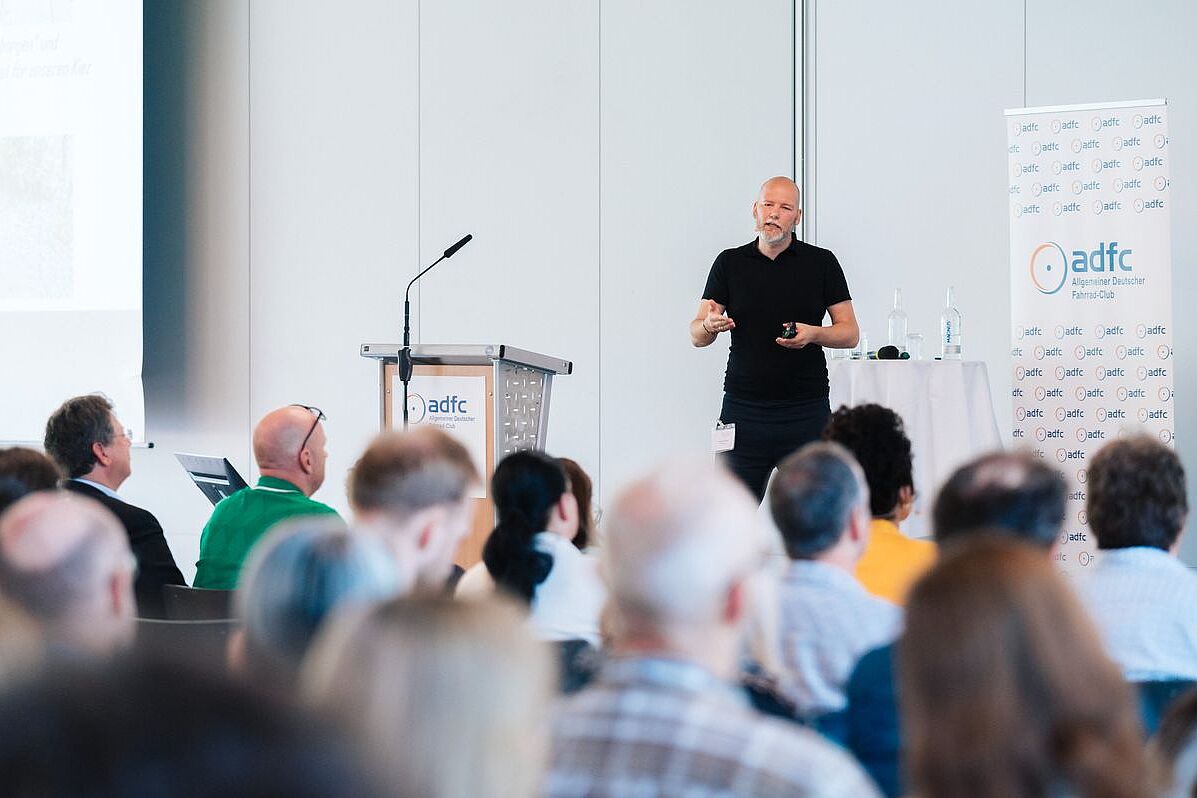 Dr. Stefan Carsten Dr. Stefan Carsten hält auf einer Bühne einen Vortrag. Im Vordergrund ist Publikum zu sehen. Im Hintergrund ein Rednerpult, weiße Wand und ein weißer Aufsteller mit ADFC-Logos.
