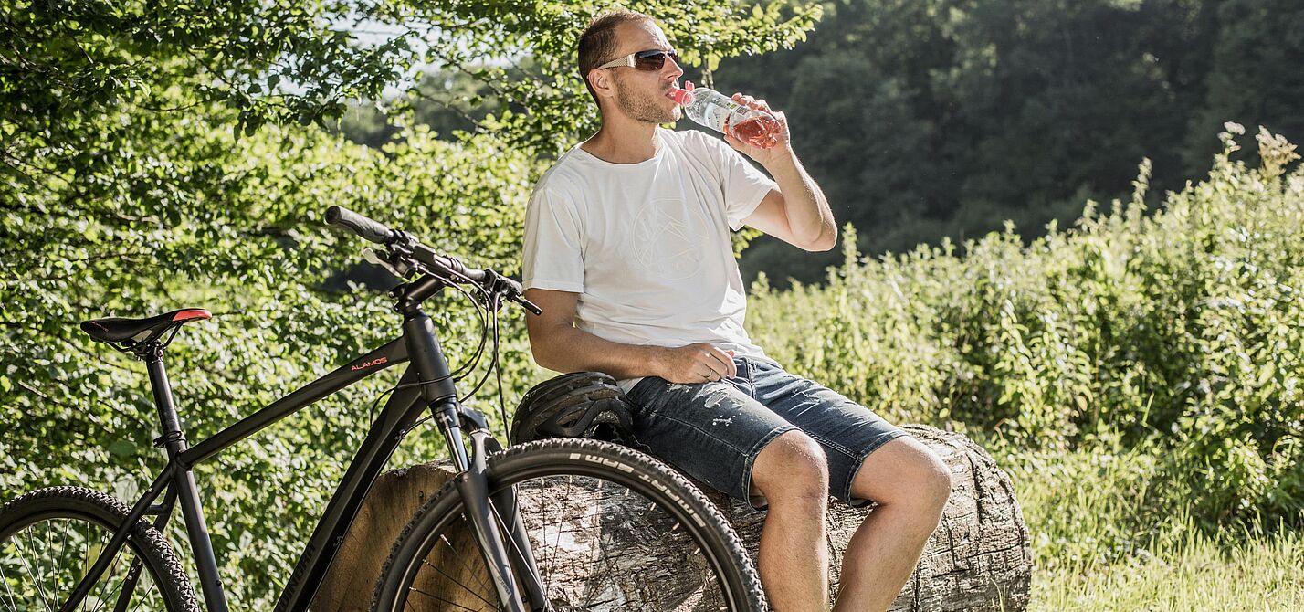 Radfahrer macht Pause im Grünen Radfahrer mit Sonnenbrille trinkt Wasser aus einer Flasche während einer Pause auf einem Baumstamm. Sein schwarzes Mountainbike steht daneben.