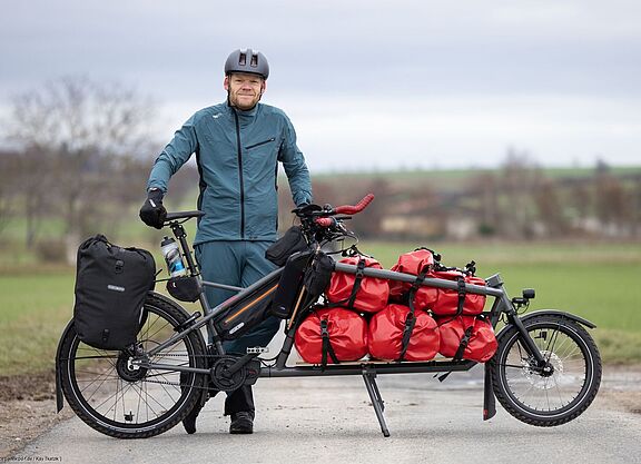 Gunnar Fehlau mit Workpacking-Lastenrad Gunnar Fehlau steht in Fahrradkleidung, vor ihm sein Lastenrad, mit dem er auf Workpacking-Tour ist.