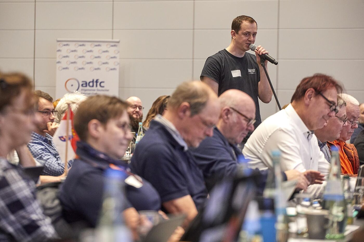 Ein Teilnehmer der BHV steht mit Mikrofon im Saal und spricht, während andere am Tisch sitzen und zuhören.
