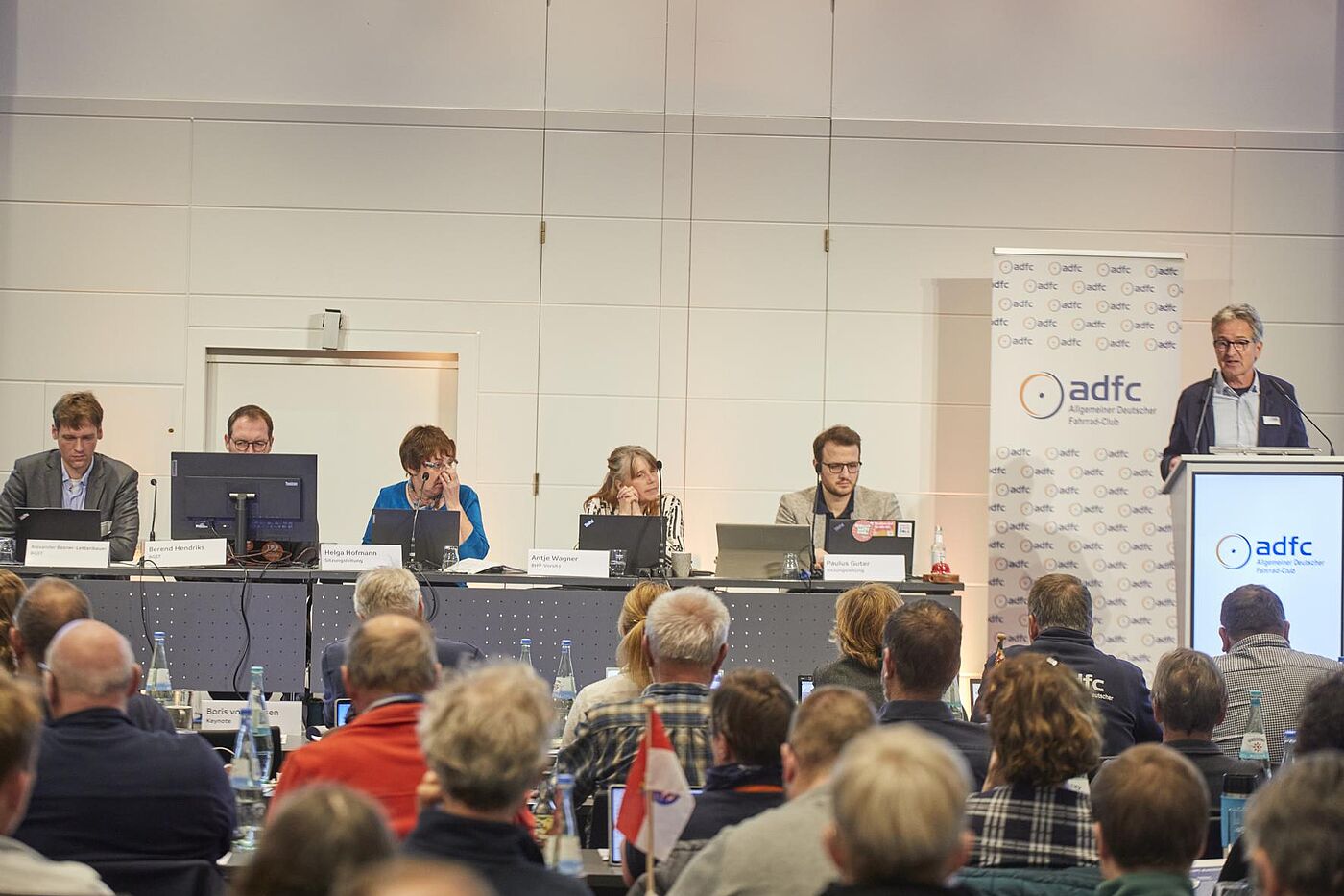 Mehrere Personen sitzen auf einem Podium vor Laptops, während rechts Christian Tänzler am Pult zu einem vollen Saal spricht.