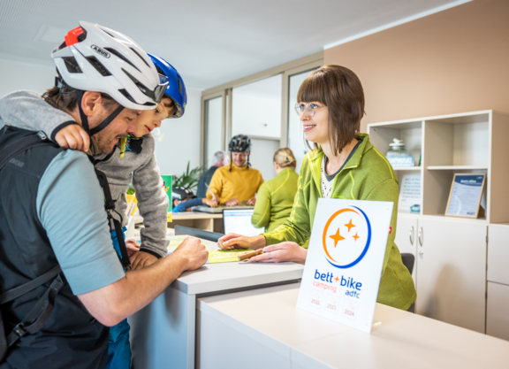 Bett+Bike - Rezeption Camping Ein Vater mit Kind auf dem Arm informiert sich an einer Rezeption eines Campingplatzes, beide tragen Fahrradhelme. Im Vordergrund steht ein Bett+Bike-Camping-Schild.