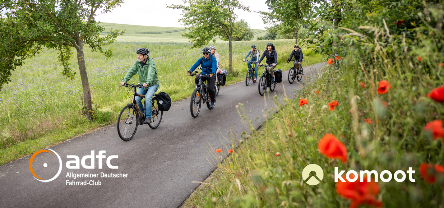 komoot Kooperation Gruppe Radfahrender auf Radweg zwischen blühenden Wiesen mit Logo ADFC und komoot
