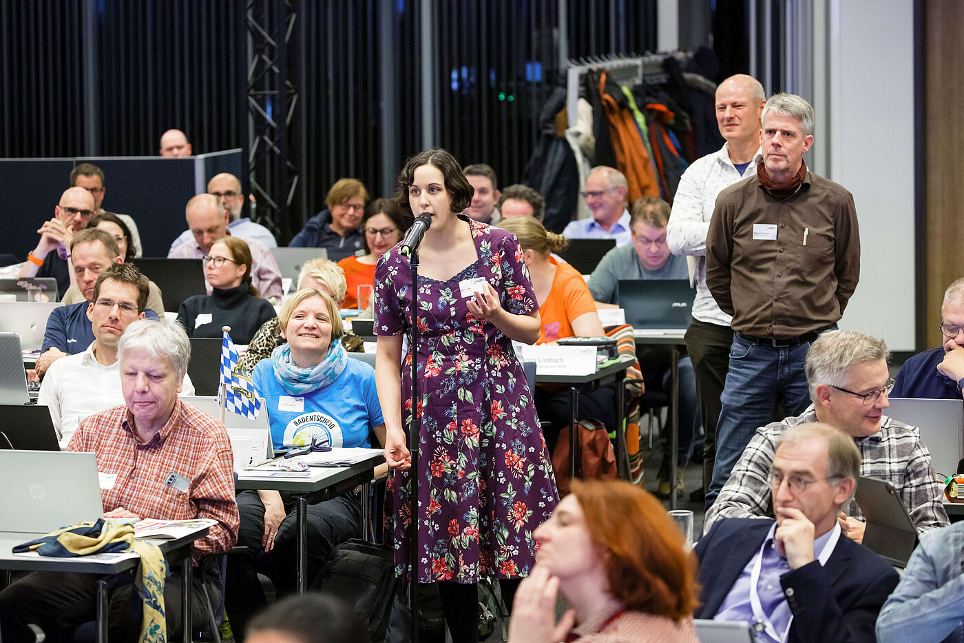 Generalaussprache Eine Frau am Mikrofon in einem Saal voller Menschen.