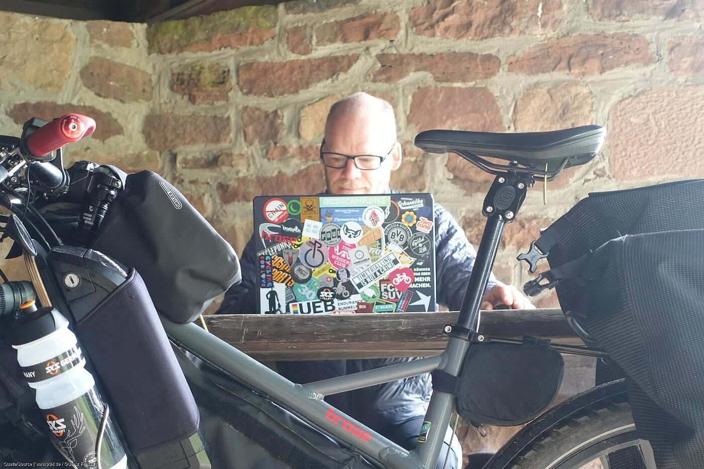 Arbeiten unterwegs. Gunnar Fehlau sitzt in einem Rasthäuschen und arbeitet am Computer. Sein Lastenrad im Vordergrund.