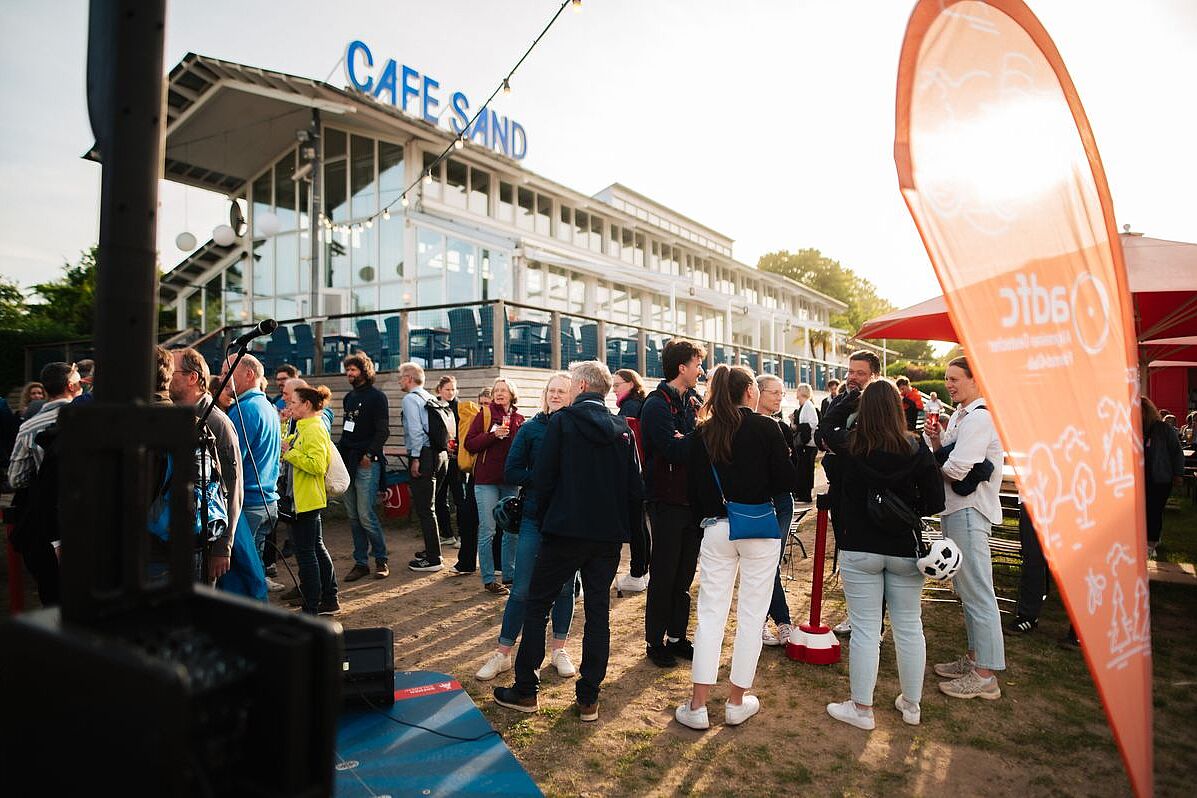 Café Sand Abendstimmung am Weserstrand. zahlreiche menschen vor dem Café Sand in Bremen in Gesprächen. Im Vordergrund eine orangefarbene ADFC-Beachflag.