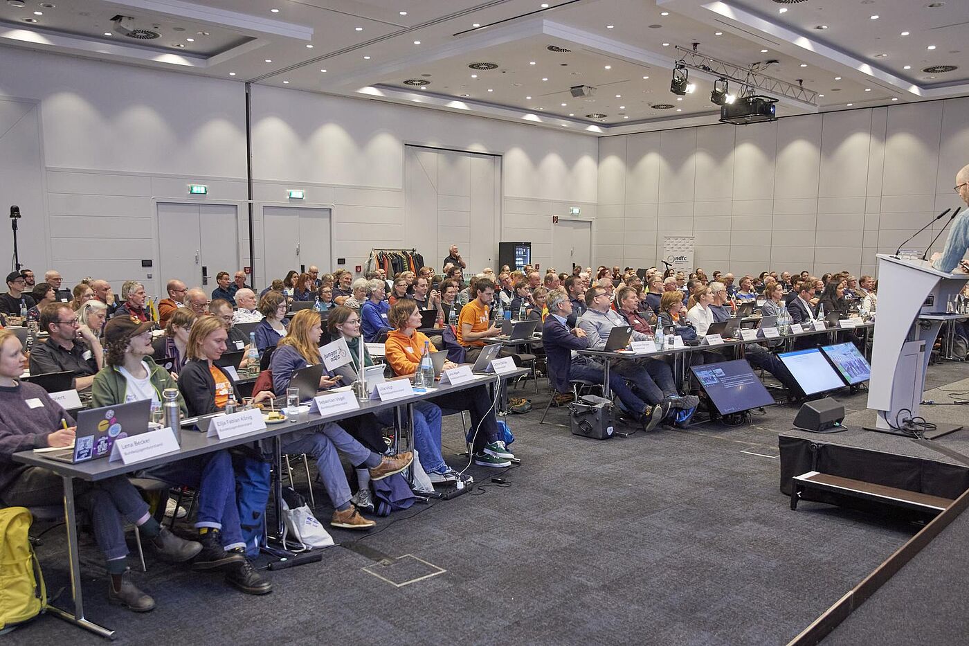 Voll besetzter Konferenzsaal mit sitzenden Delegierten, die einem Redner am Pult folgen.