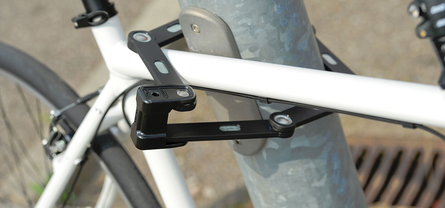 Anschließen statt nur abschließen! weißes Fahrrad ist mit einem Faltschloss an eine Metallstange angeschlossen