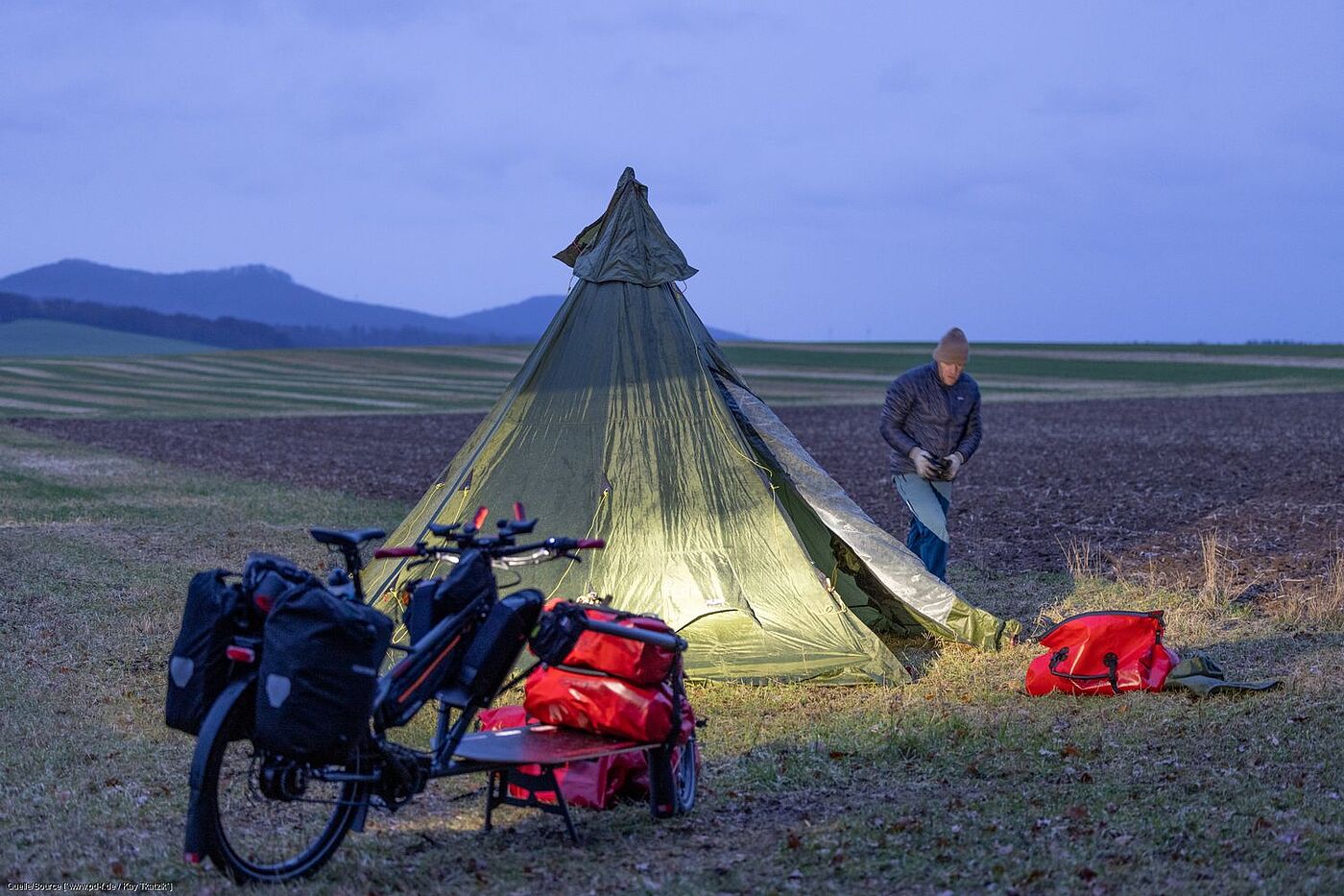 Workpacking: Übernachten im Zelt Ein Zelt steht auf einem Feld, davor ein Lastenrad. Gunnar Fehlau läuft um das Zelt herum, um Heringe zu befestigen.