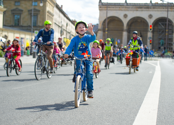 Kinder fahren mit dem Fahrrad bei einer Familien-Fahrraddemo. Ein Kind im Vordergrund winkt in die Kamera.